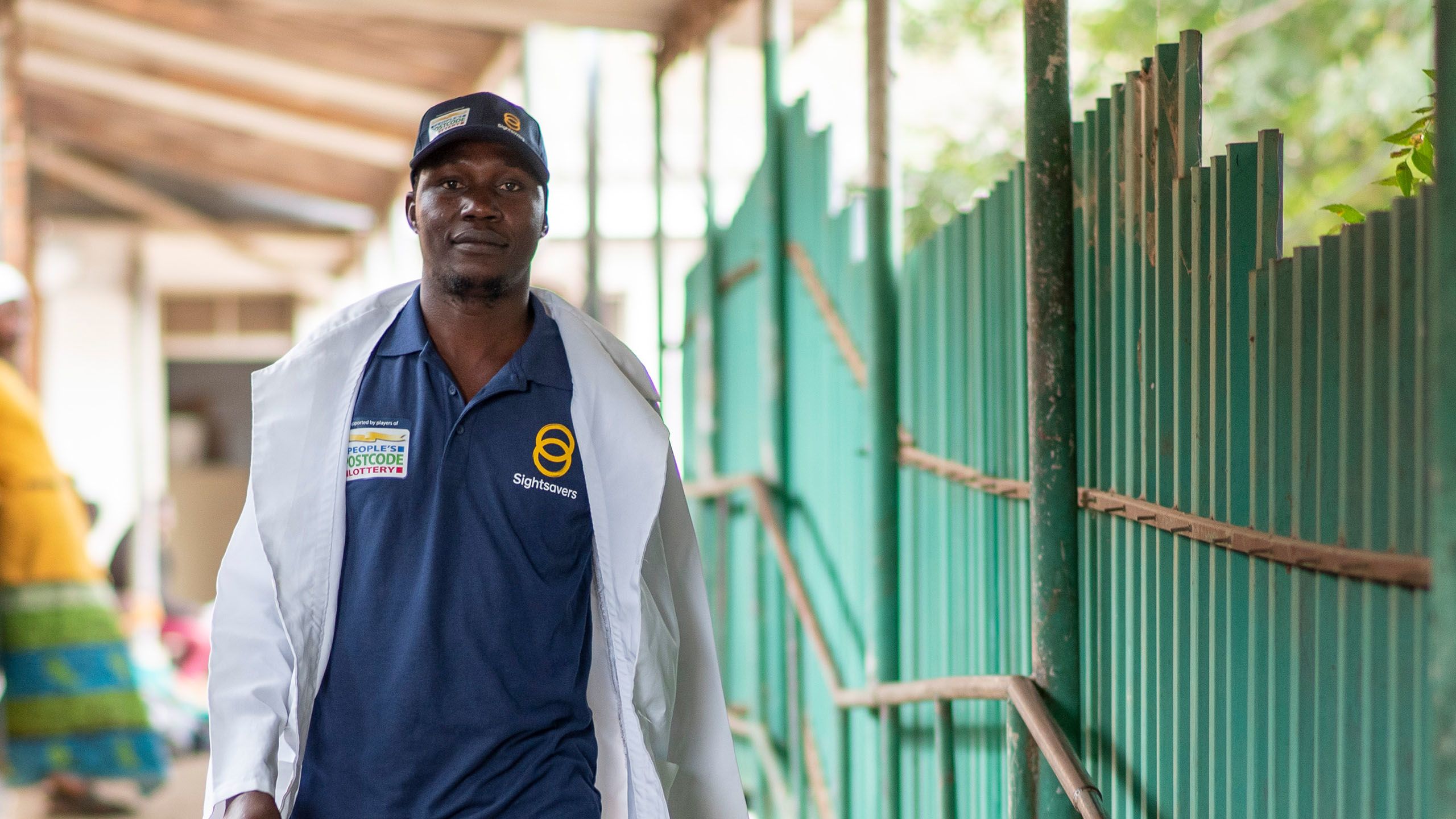 Optometrist George Richards walking confidently towards the camera, looking into the lens, wearing a white lab coat open at the front and a Sightsavers and People's Postcode Lottery branded navy blue polo shirt and baseball cap.