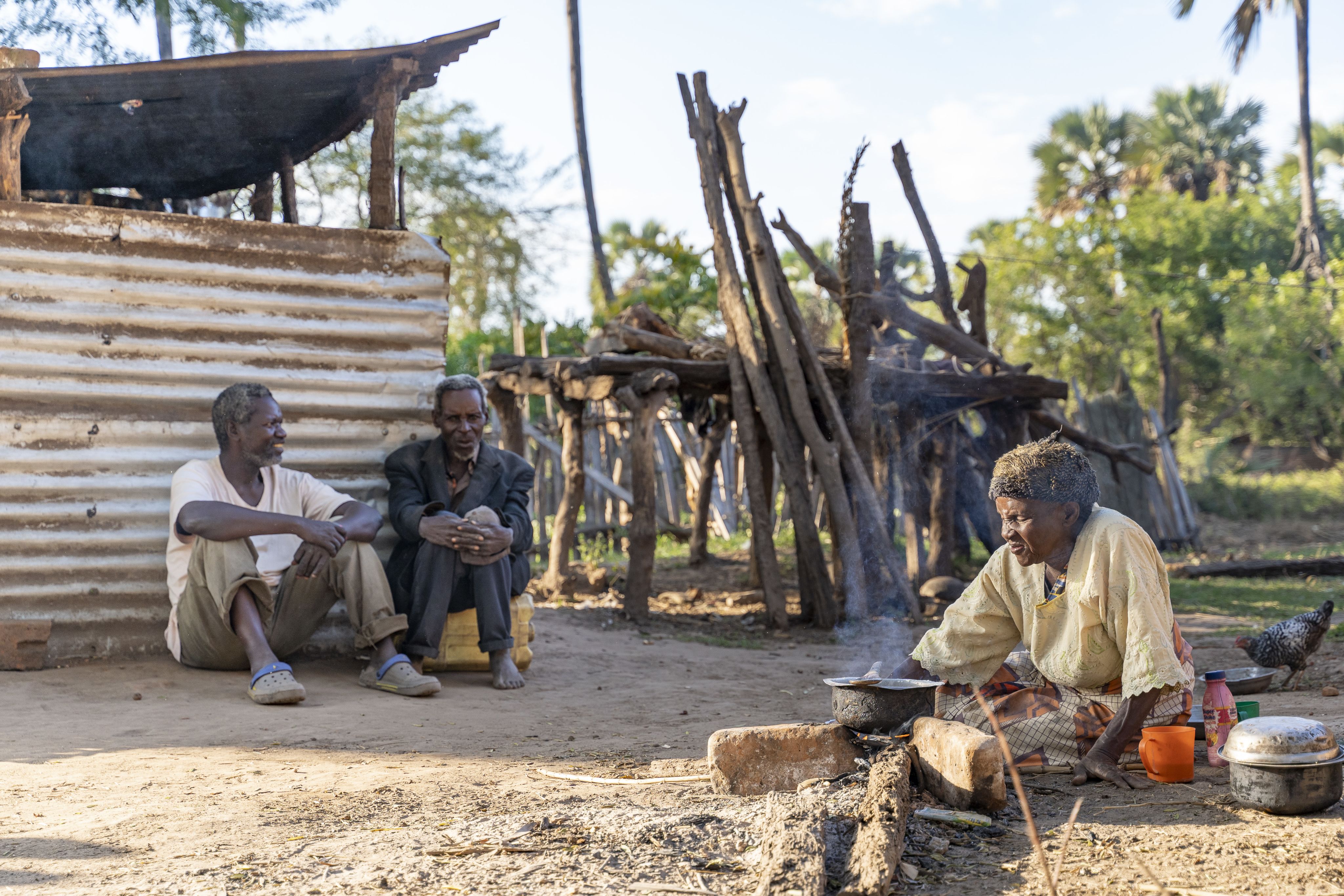 Wilson, Ganizani and Alice sit outside in the village having a discussion. Alice is preparing food in a pot over an open fire.